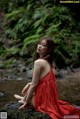 A woman in a red dress sitting on a rock by a stream.